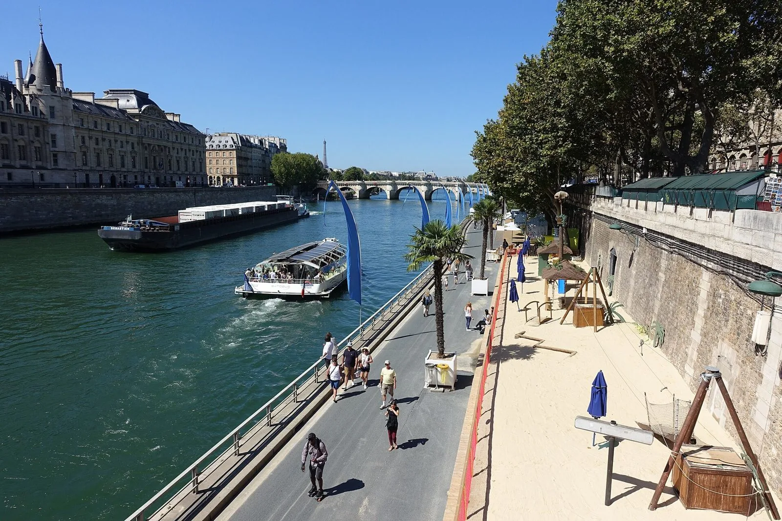 Lungosenna soleggiato d'estate con palme in vaso, sabbia e un battello sul fiume, sullo sfondo la Conciergerie e il Pont Neuf